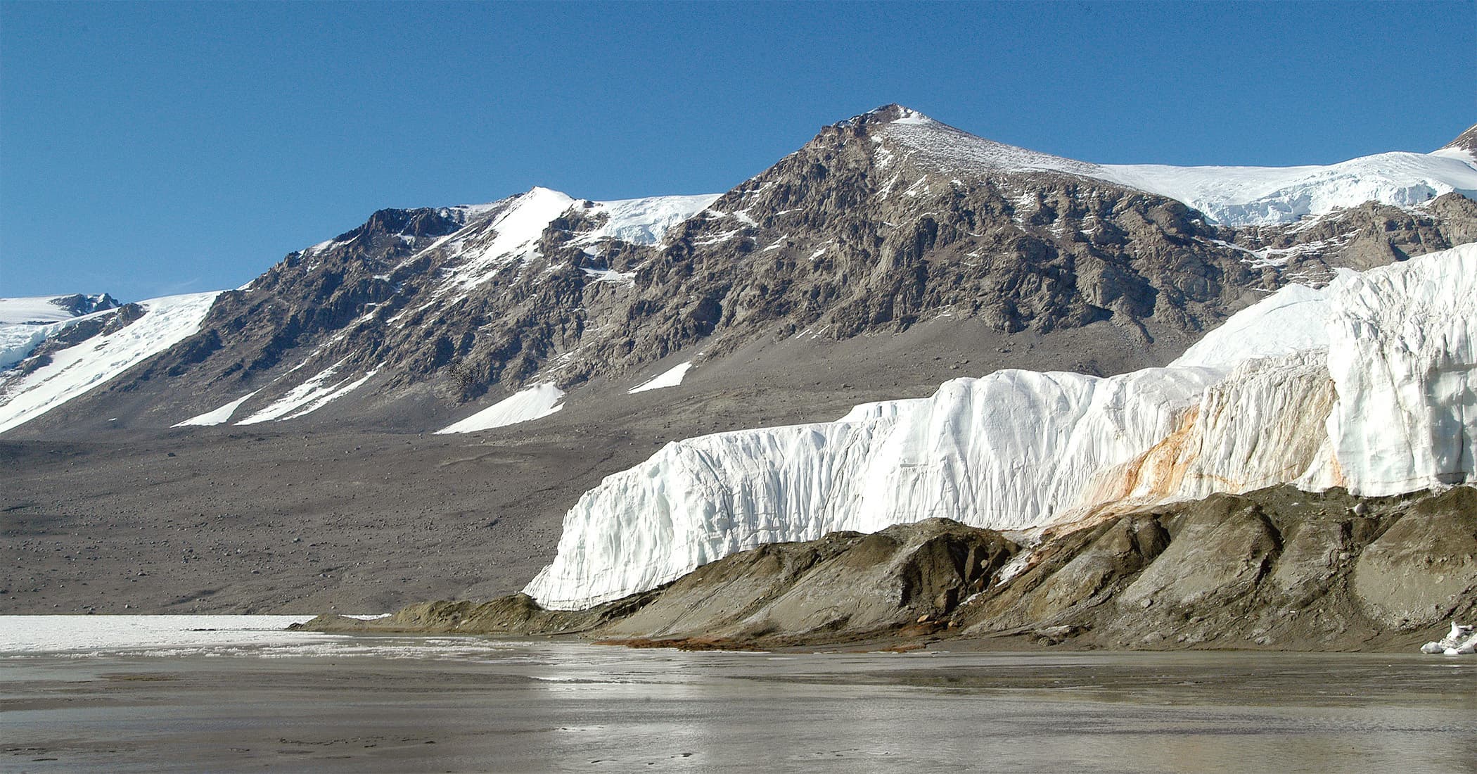 テイラー氷河の実写。右側の白い氷壁に赤橙色の跡があり、手前には湖、後ろには乾いた岩山が見える